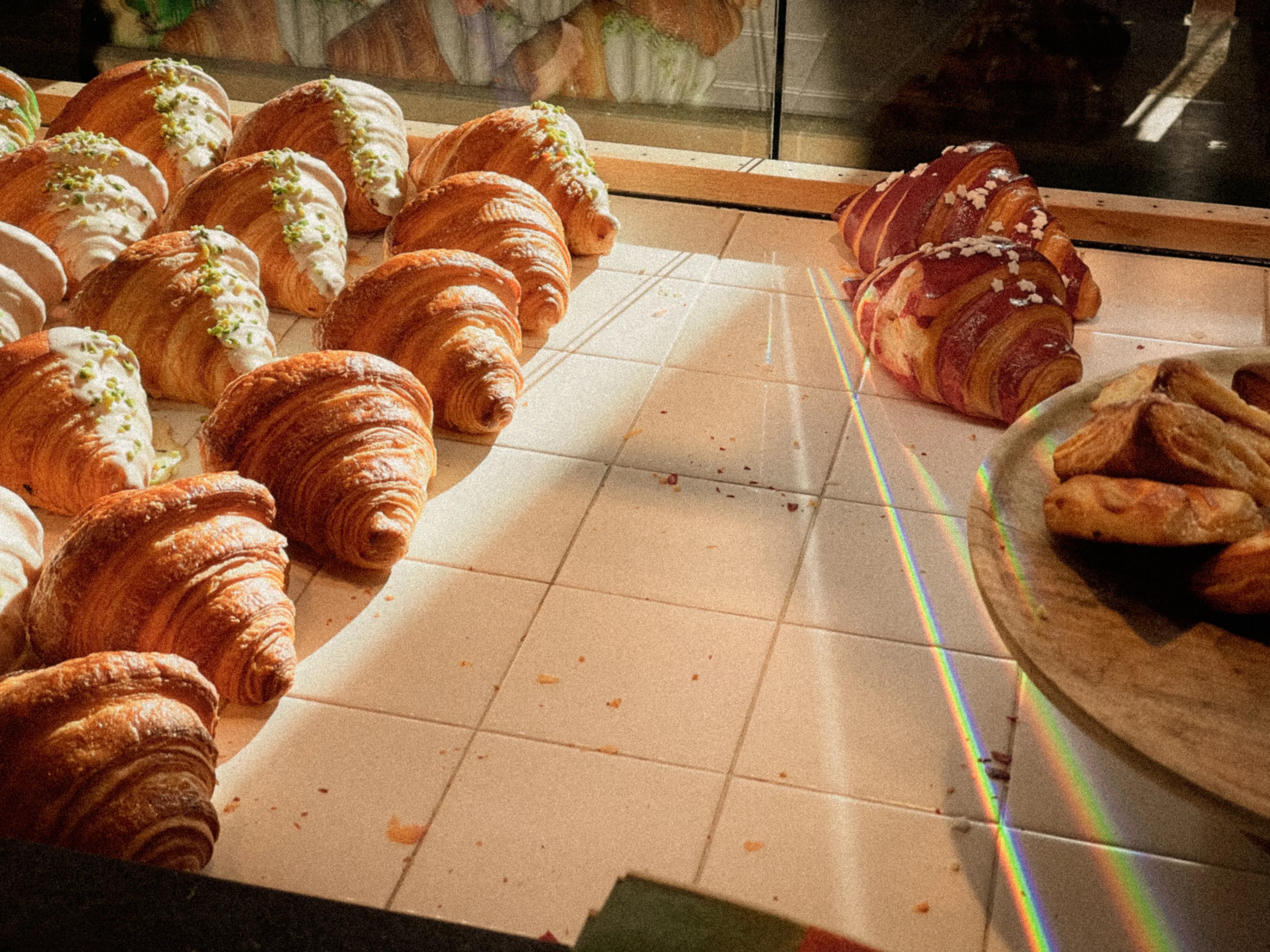 Rows of croissants and pastries lit by sunlight across a tiled counter
