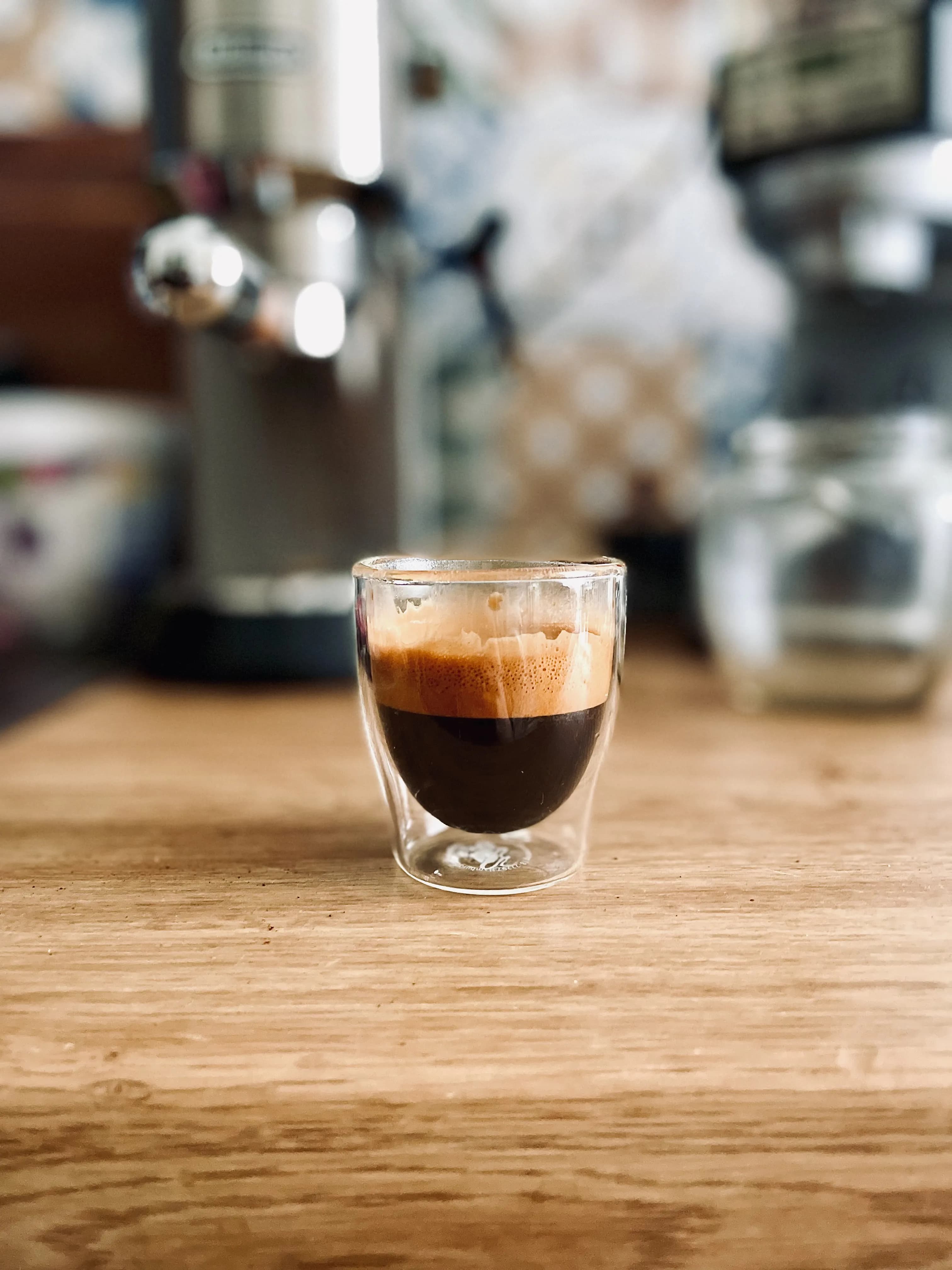Small glass of espresso on a wooden counter with a coffee machine behind it